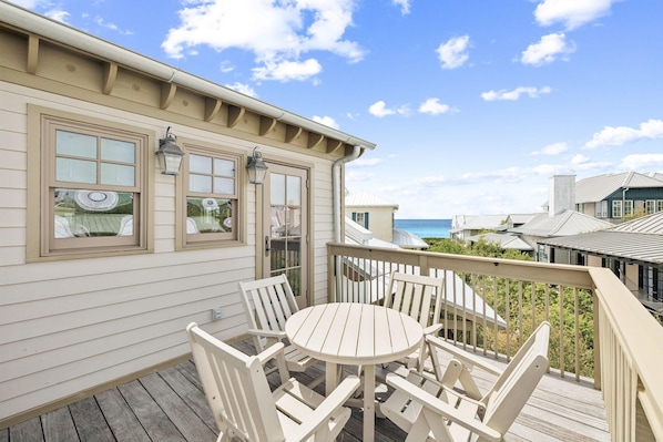 A deck with a round table and chairs, a railing, and a view of the ocean.