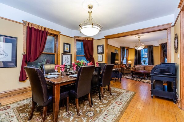 Un coin repas avec une table en bois dressée pour quatre personnes, entourée de chaises sombres. Un lustre est suspendu au plafond, et une cheminée est visible au fond.