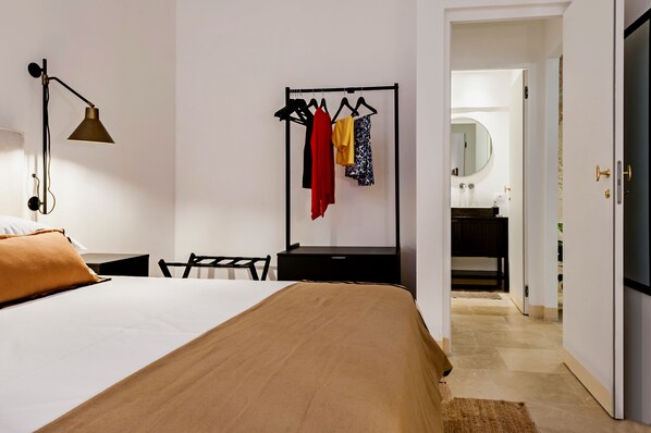 A hotel room with a bed, a desk, a chair, a hanging clothes rack, and a bathroom visible through an open door.