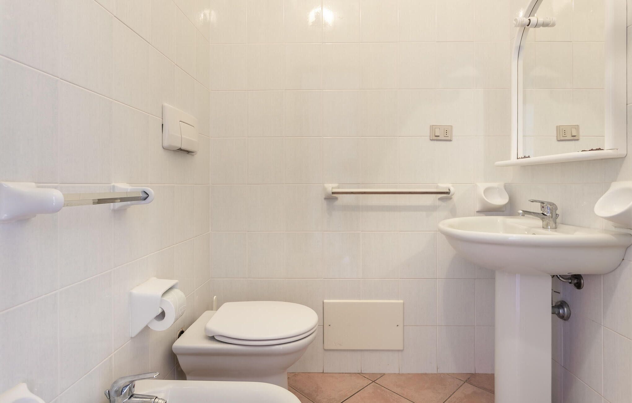 Et moderne badeværelse med hvide fliser, toilet, bidet, vask og spejl.