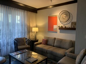 Un salon comprenant un canapé, un fauteuil, une table basse et une lampe. Il y a une fenêtre avec des rideaux et un mur orné d’un élément décoratif.