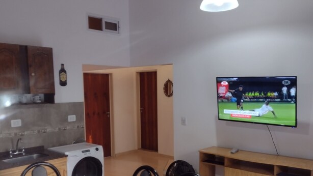 Una zona de menjador amb una taula de vidre i cadires, una cuina amb rentadora, armaris de fusta i un televisor que mostra un esdeveniment esportiu.