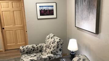 A living room with a patterned armchair, a small table with a lamp, a framed picture on the wall, and a wooden door.