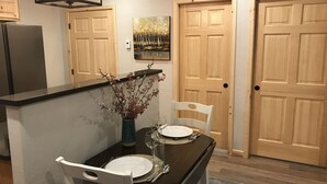Ein Essbereich mit einem dunklen Holztisch, gedeckt für zwei Personen, weißen Stühlen, einer Vase mit getrockneten Blumen und einem Bild an der Wand.