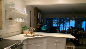A modern kitchen with white cabinetry, a stainless steel range, and a built-in oven. There is a sink with a gold faucet, and a small island with a plant. The kitchen opens to a dining area with a chandelier and a view of trees through large windows. - Bluebird Rest - Peaceful Lake Retreat (Kelso)