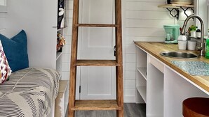 Ett sovrum i loftstil med en trästege, en säng med mönstrat sänglinne, en liten köksdel med diskho och hyllor samt ett fönster med utsikt.