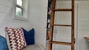 Ett rum med en säng på ett loft, en trästege, ett fönster och en vit dörr.