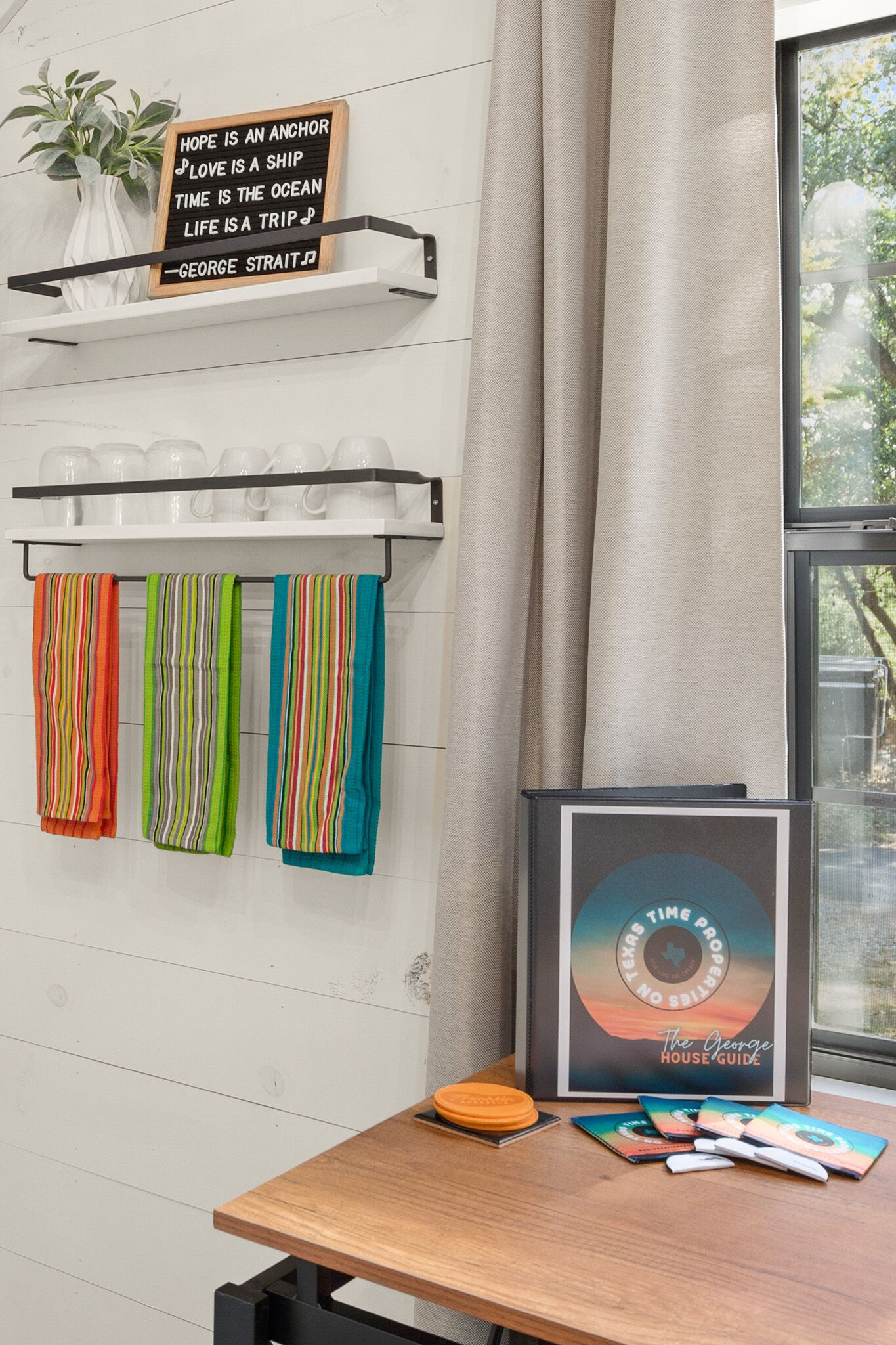 Una habitación con pared blanca, una estantería con un letrero, una ventana con cortinas y una mesa con un cuadro enmarco y libros.