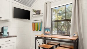 Una cocina con una mesa de madera, taburetes de bar, un televisor y un ventanal con cortinas.