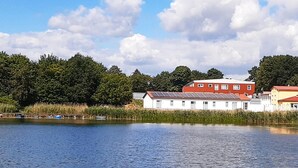 A lakeside view with a row of white buildings, some with solar panels, and dense green trees in the background. - 2 bedroom nice home in Gützkow (Gützkow)