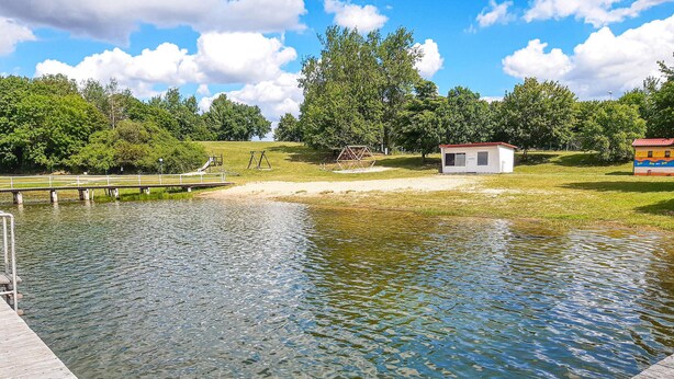 Un parque tranquilo con un muelle de madera que se adentra en un lago sereno, rodeado de abundante vegetación y un cielo azul despejado.