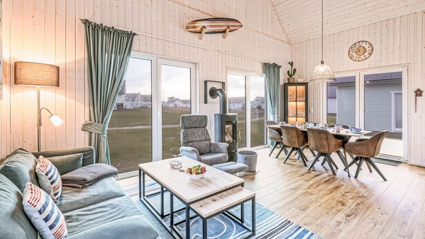 Een gezellige woonkamer met een bank, een salontafel en een open haard. Er is een eethoek met een tafel en stoelen. De kamer heeft houten wanden en een groot raam.