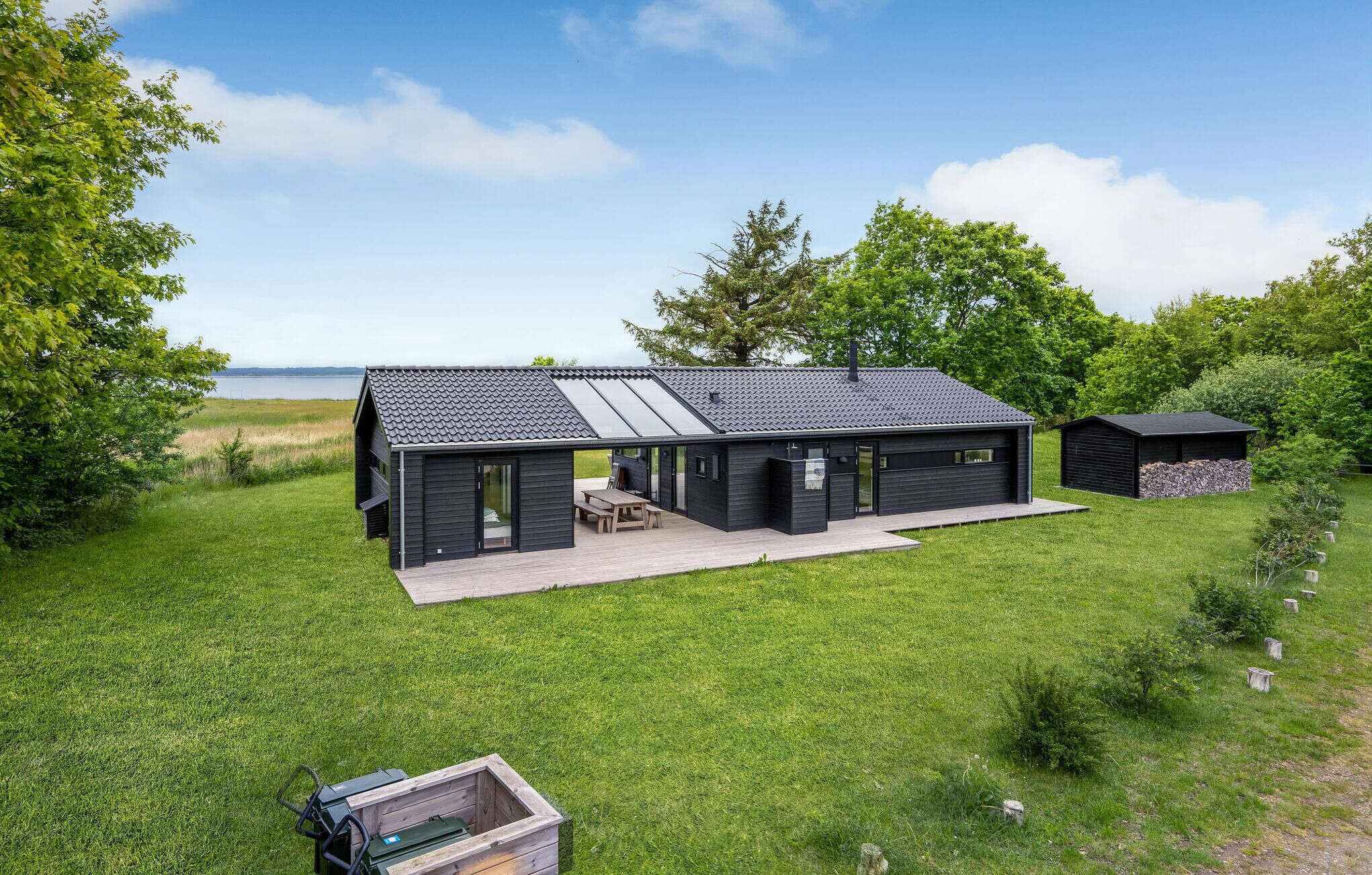 A modern cabin with a large deck, a garage, and a small shed, surrounded by lush greenery and a lake view.