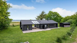 A modern cabin with a large deck, a garage, and a small shed, surrounded by lush greenery and a lake view.