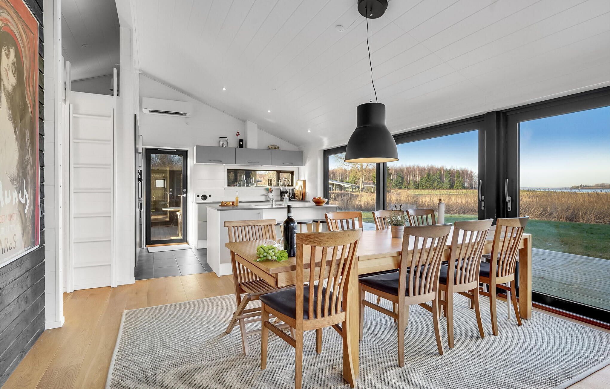 A modern kitchen with a large island, a dining area with wooden chairs, and a view of a lake through large windows.