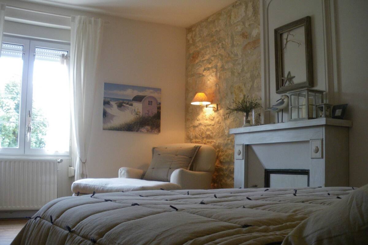 A bedroom with a bed, a chair, a fireplace, a painting, and a window.