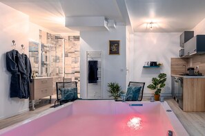 Une salle de bain moderne avec une douche à parois de verre, une baignoire éclairée d’une lumière rose et un petit coin cuisine équipé d’un évier et de placards.