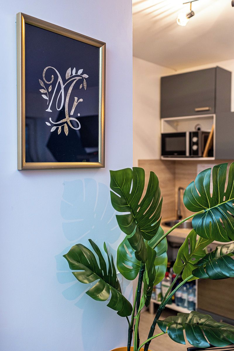 Een lijst met een monogram aan een muur, een pot met een monstera plant, en een keuken op de achtergrond.