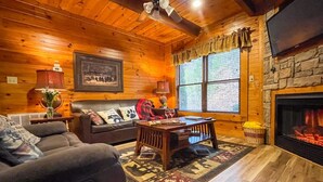 Un intérieur de chalet chaleureux avec une cheminée, des murs en bois, un mur d’accent en pierre, un ventilateur de plafond et une grande fenêtre.