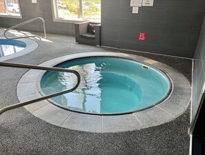 Una piscina interior pequeña con una escalera metálica, paredes de azulejos y una ventana con vista a coches estacionados.