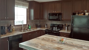 A modern kitchen with dark wooden cabinets, a granite countertop, a built-in microwave, a stainless steel sink, a dishwasher, and a refrigerator. - Rica Rico - Beach View Getaway (Grand Isle)