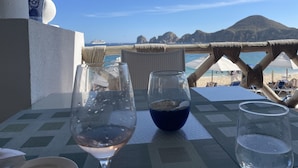 A table with two glasses, one with a blue drink and another with a clear liquid, overlooking a beach with a mountain in the background. - Rose is located directly on Medano Beach the best swimmable beach in Los Cabos (Cabo San Lucas)