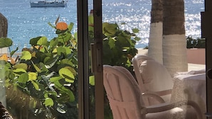 A view through a glass door of a room with a chair, a palm tree, and a boat on the water. - Rose is located directly on Medano Beach the best swimmable beach in Los Cabos (Cabo San Lucas)