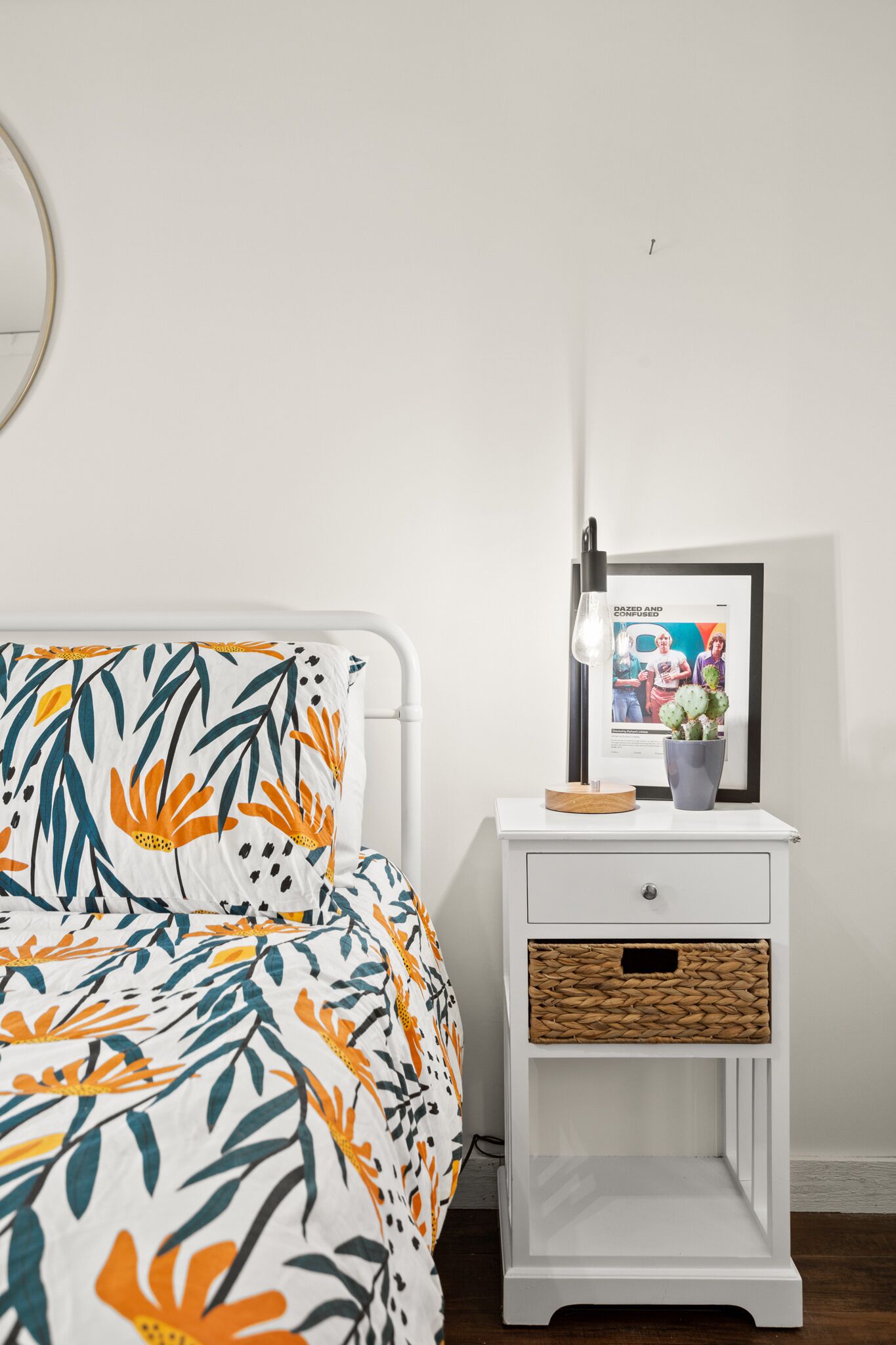 Una cama bien hecha con una colcha estampada con motivos florales, una mesita de noche con una lámpara, un cuadro enmarcado y una pequeña planta en maceta.