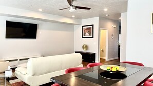 Un salon moderne avec un canapé blanc, une table à manger avec des chaises rouges, un ventilateur de plafond et un téléviseur fixé au mur.