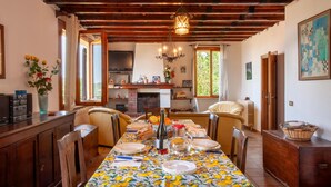 Comedor con una mesa larga dispuesta para seis, vigas de madera en el techo, una araña y una cocina al fondo.