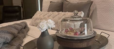 Een netjes opgemaakt bed met een decoratief dienblad, een vaas met witte bloemen en een glazen stolp met chocolade.