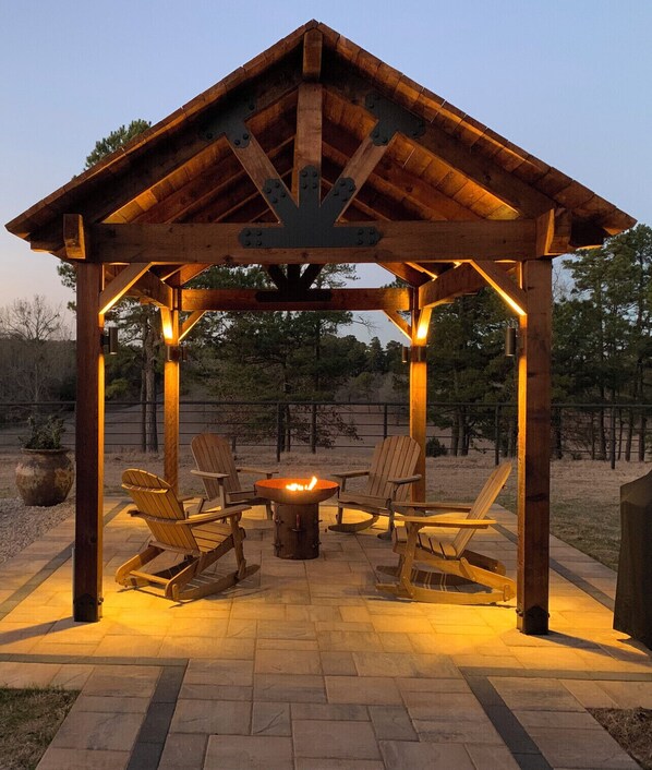 Relax in a serene gazebo with a central fire pit, surrounded by Adirondack chairs on a paved patio.