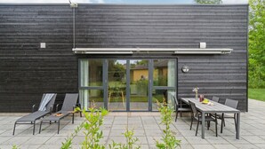 Ein modernes Gebäude mit dunkler Fassade, großen Schiebeglastüren, Sitzgelegenheiten mit Stühlen und Tischen im Freien und einer kleinen Grünfläche im Vordergrund.
