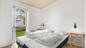 Ein Schlafzimmer mit einem Bett, zwei Nachttischen, einem Fenster mit Blick auf Bäume und einer Deckenleuchte.