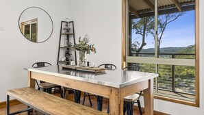 Un espace repas avec une table en bois, des bancs et des chaises. Une grande fenêtre offre une vue sur la verdure et les arbres.