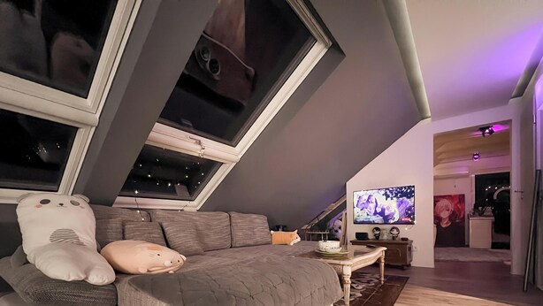 Un dormitorio tipo loft acogedor con techo inclinado, un ventanal grande, una cama con colcha gris, un almohadón blanco mullido y un televisor sobre un mueble.