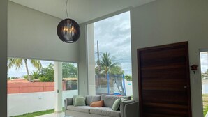 A modern living room with a large window, a sofa, and a wooden door. - Barra do Jacuípe - House with 3 bedrooms, 1 of which is a suite. (Jacuípe)