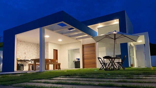 A modern house with a patio area, a dining table, and a large umbrella. - Barra do Jacuípe - House with 3 bedrooms, 1 of which is a suite. (Jacuípe)