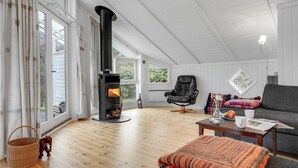 Ett mysigt vardagsrum med en vedeldad kamin, en fåtölj i läder, en soffa och ett matbord med en vas och en tidning.