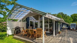 En täckt uteplats med trädgårdsmöbler och en grill, omgiven av ett trädäck och en inglasad konstruktion.