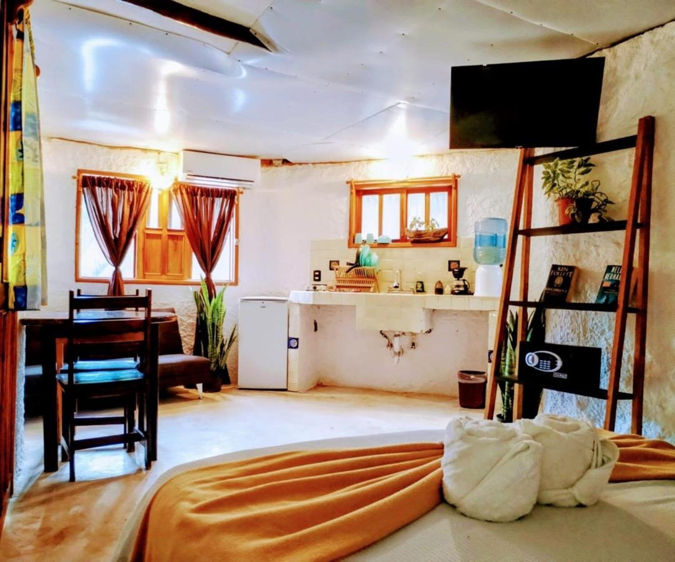 A compact room with a bed, a desk, a chair, a small kitchenette, and a TV mounted on the wall.