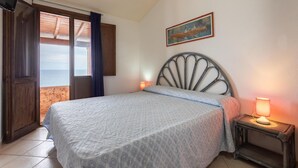Une chambre à coucher avec un lit, une table de chevet avec une lampe, un tableau au mur et une vue sur la mer par la fenêtre.