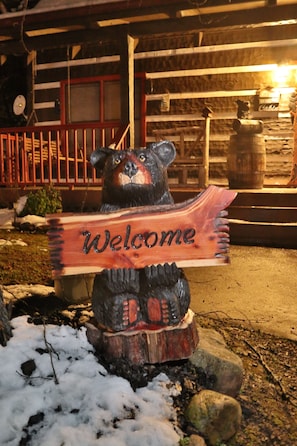 A wooden bear holding a 'Welcome' sign, set against a backdrop of a rustic building with a red railing and a stone wall.
