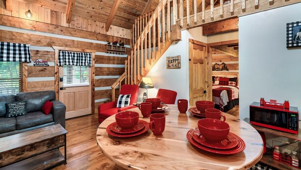 Ein gemütliches Blockhaus-Interieur mit Holzbalkendecke, einem Esstisch mit roten Geschirr, einem roten Sofa und einem roten Sessel.