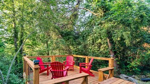 Eine Holzterrasse mit roten Adirondack-Stühlen und einem Picknicktisch, umgeben von dichtem Wald.
