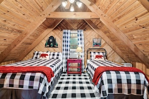 A cozy attic bedroom with two beds, checkered bedding, a red nightstand, and a window with plaid curtains.