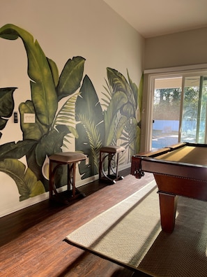 A pool table in a room with a large mural of tropical leaves on the wall, wooden flooring, and a window with a view of trees.