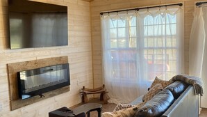 A cozy living room with a wooden ceiling, a flat-screen TV mounted on the wall, a leather sofa, a coffee table, and a large window with sheer curtains. - One room cabin set on our farm for a peaceful getaway. (Union Hall)