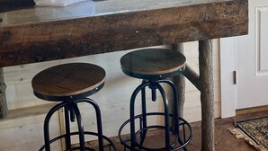 A rustic kitchen with a large wooden table, two stools, and a window with a view of buildings outside. - One room cabin set on our farm for a peaceful getaway. (Union Hall)