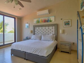 A bedroom with a bed, a ceiling fan, an air conditioning unit, and a view of palm trees through a large window.